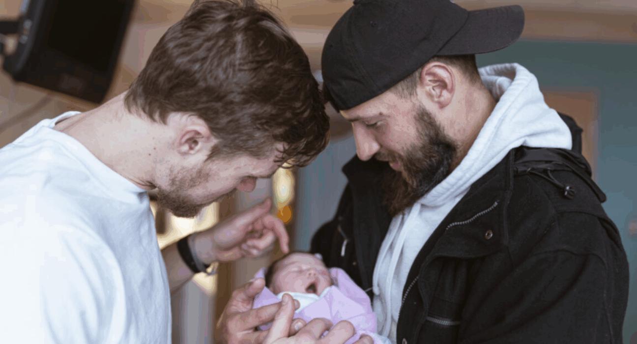Two men holding a baby wrapped in a pink blanket.