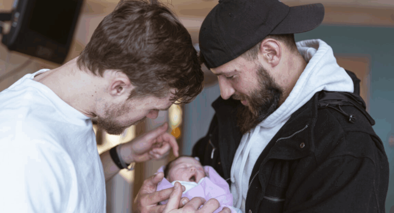 Two men holding a baby wrapped in a pink blanket.