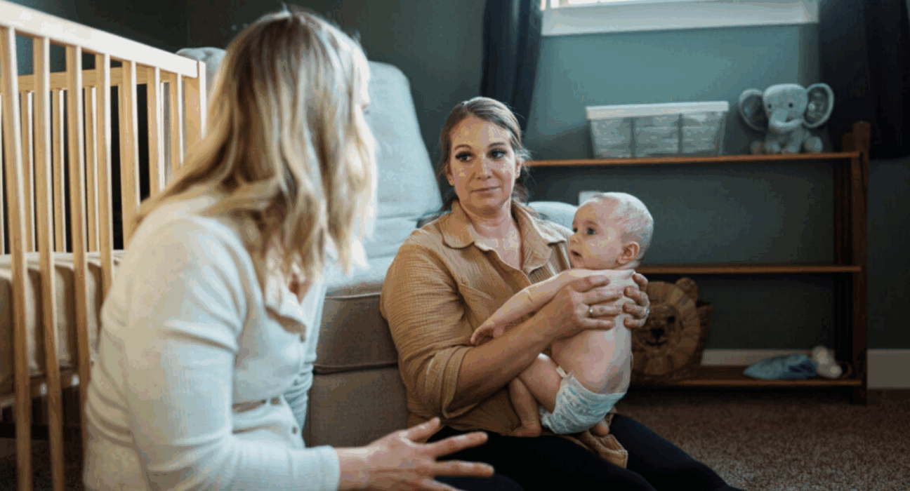 Parent holding her baby who is wearing a nappy, sitting on the floor talking to another lady, who is leaning on the cot.