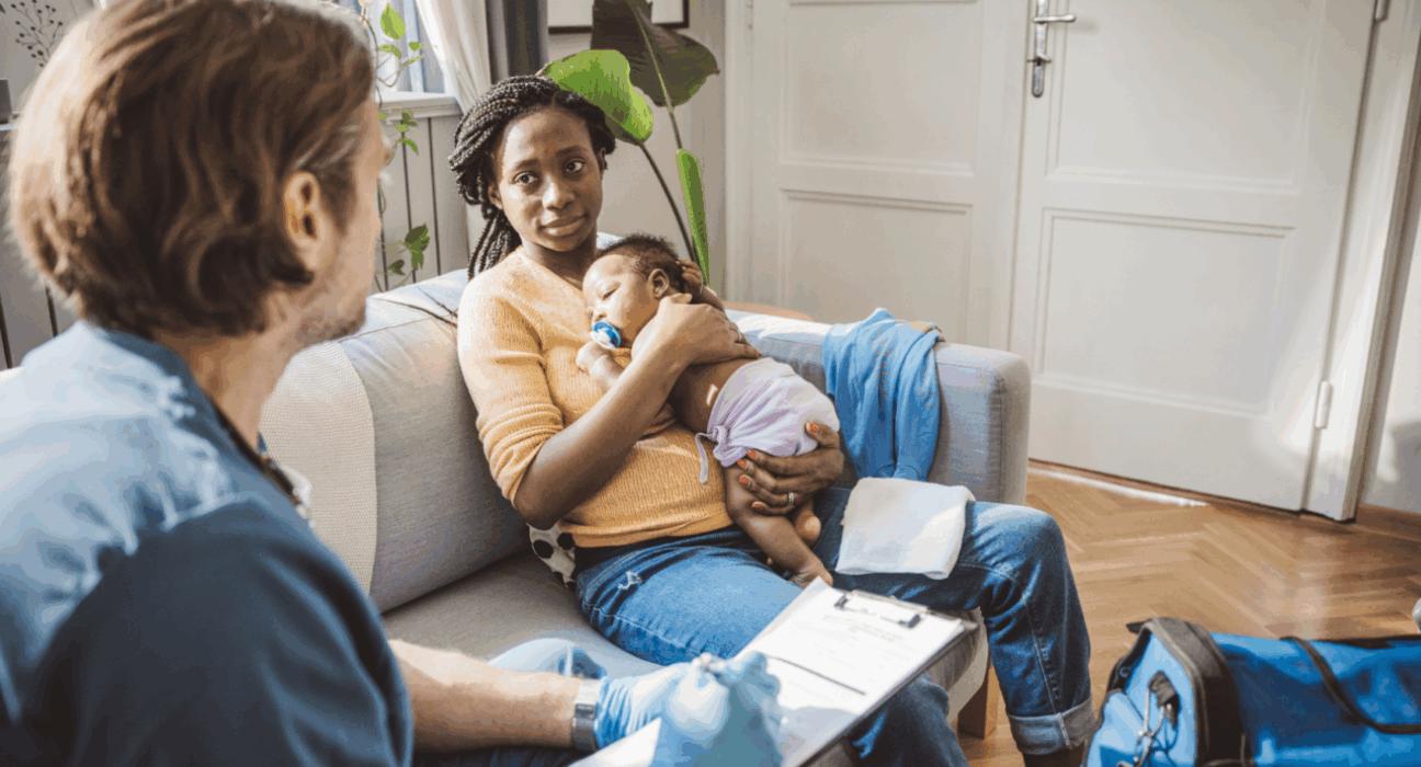 Lady holding her baby, sitting on the sofa talking to a health professional.