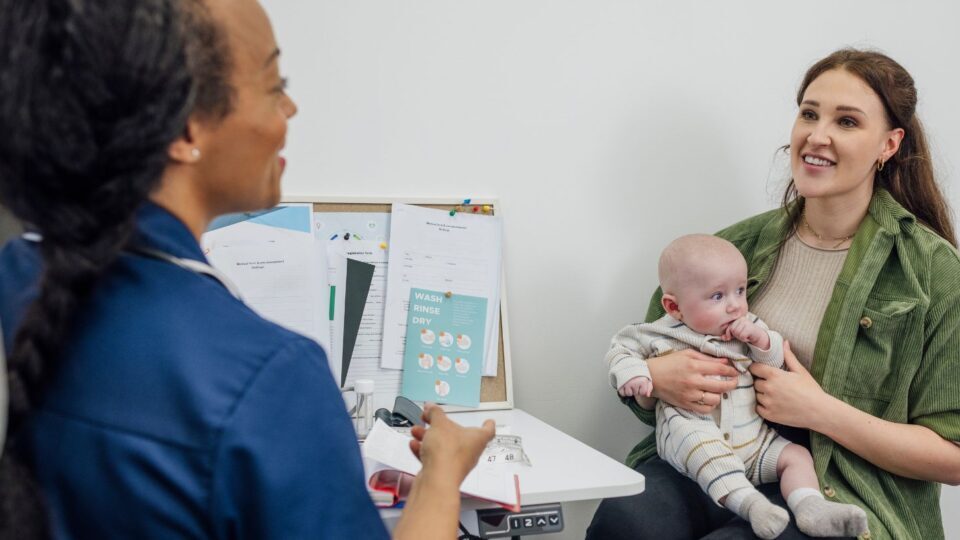 A mother is talking to a nurse at an appointment. They are smiling and the mother holds a young baby in her arms.