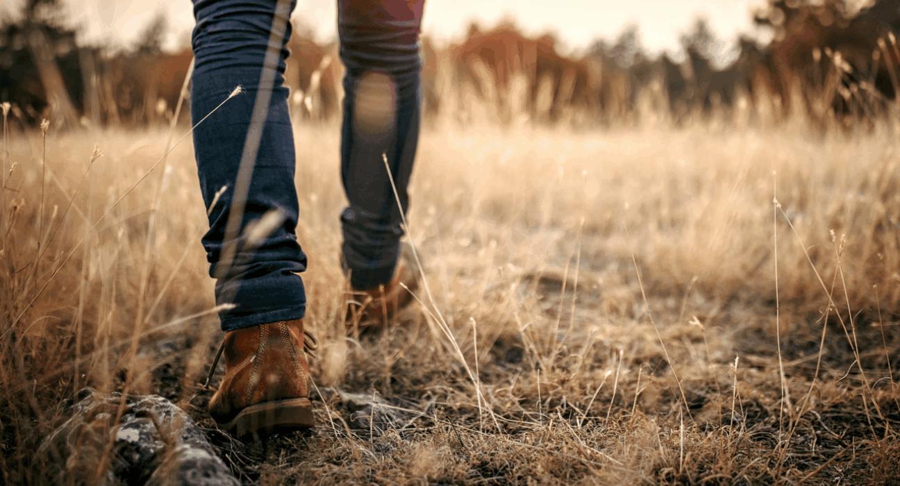 A man's legs and shoes walk through a field with long and short grass. It looks dry and the sun seems to be setting in the background.