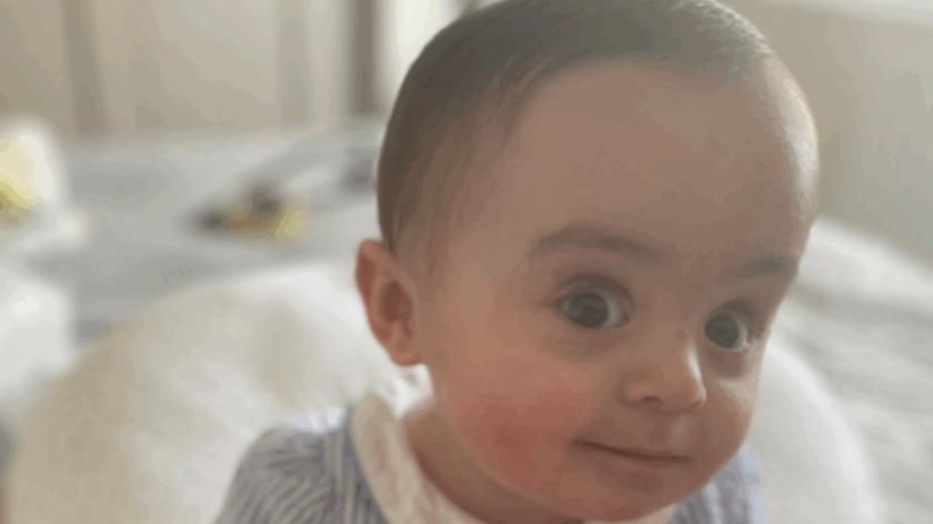 Baby Gigi sitting on a bed wearing a blue dress, looking at the camera.