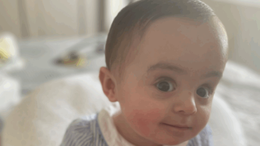 Baby Gigi sitting on a bed wearing a blue dress, looking at the camera.