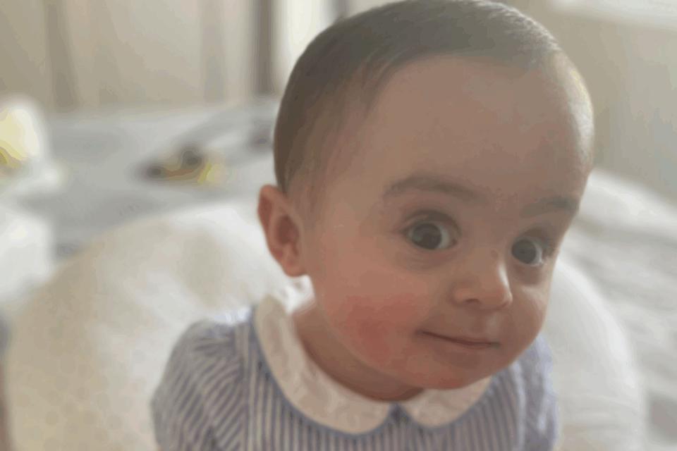 Baby Gigi sitting on a bed wearing a blue dress, looking at the camera.