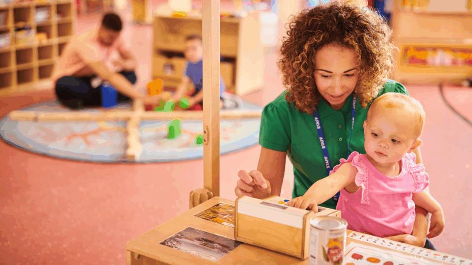 Early years professional sitting on the floor in a nursery with a little girl on her lap, playing with toys.