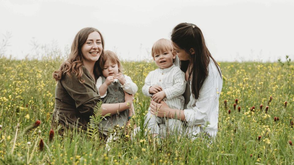 Izzy and Laura, each holding Alo and Jovie, standing in a field with tall grass.