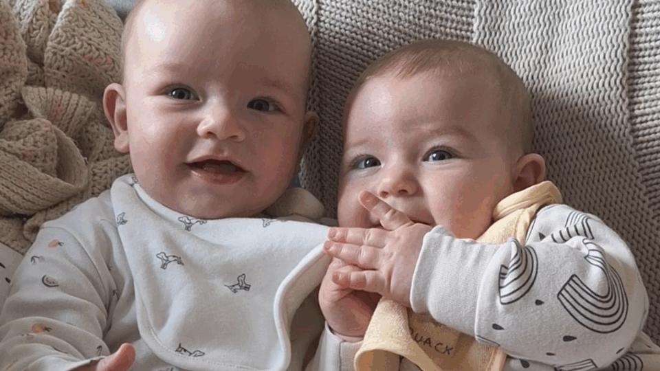 Alo and Jovie lying awake next to each other, wearing white baby grows.