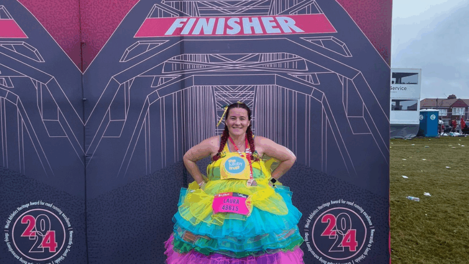 Laura wearing a colourful pink tutu and a Lullaby Trust running best, standing in front of a finisher backdrop with her hands on her hips.