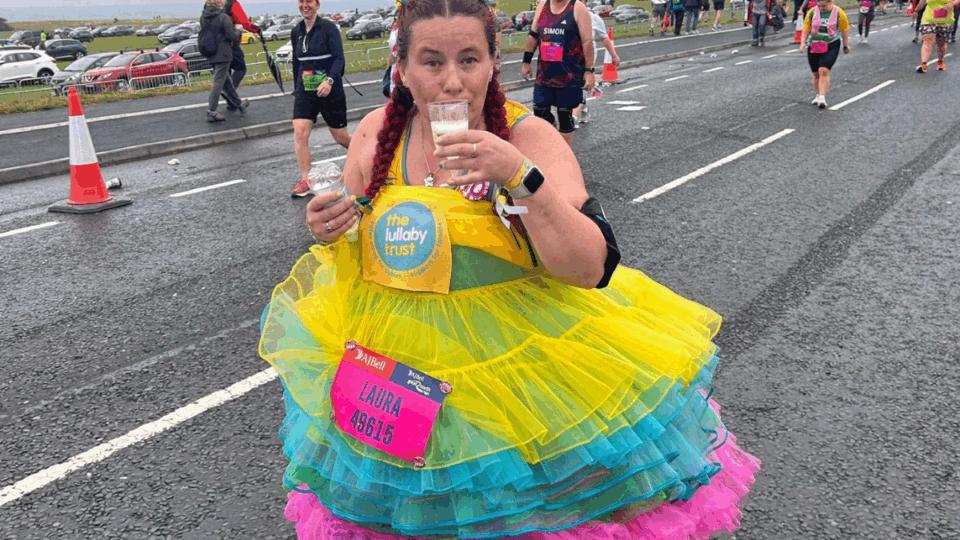 Laura wearing a big colourful tutu, sipping a drink whilst running at an event. She has a Lullaby Trust running vest on.