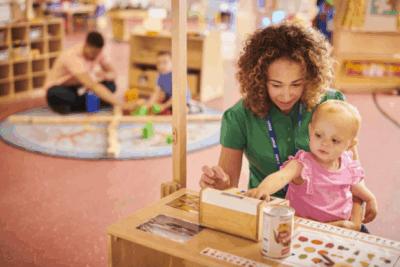 Early years professional sitting on the floor in a nursery with a little girl on her lap, playing with toys.