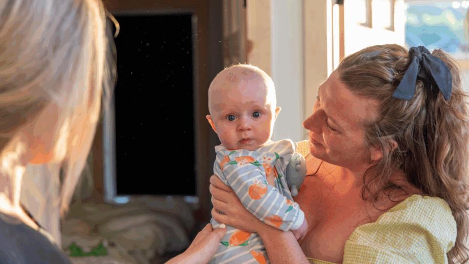 Lady holding a baby, and another lady standing in front of her with her hand on the baby's back.