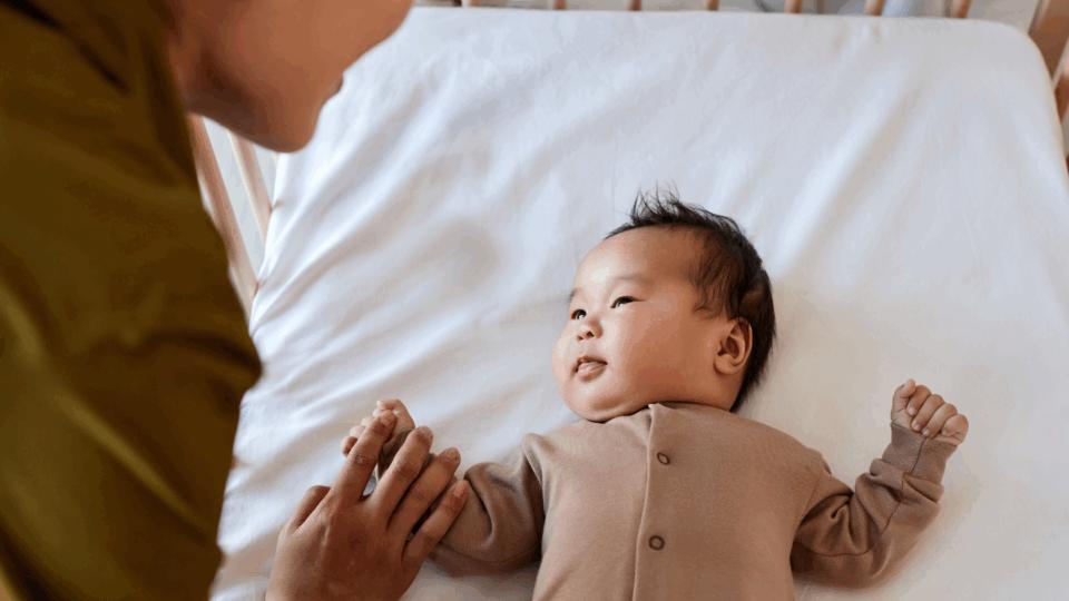 A baby is being put down to sleep in a cot. They are looking up at their parent.