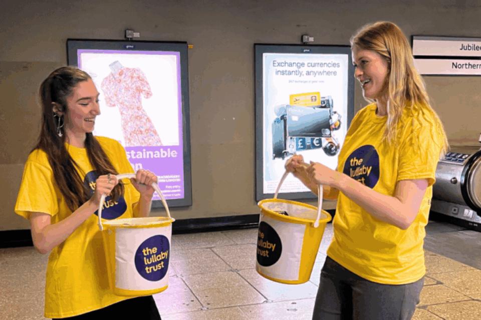 Two young women in Lullaby Trust t-shirts hold buckets to collect money in a London Underground station