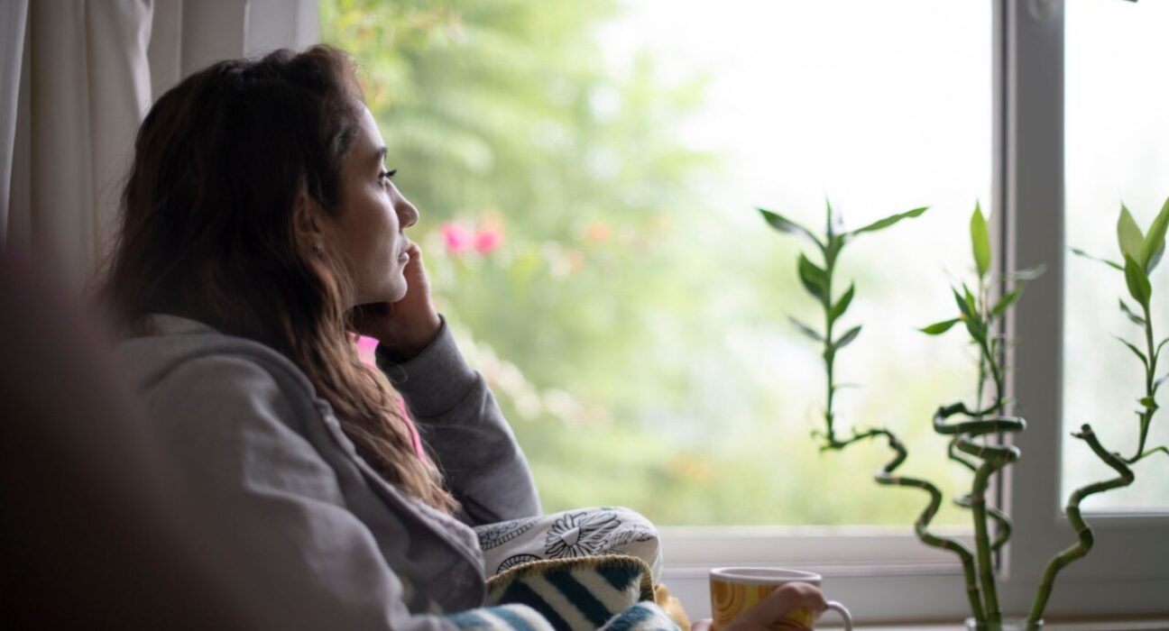 A woman is resting her chin on her hand, looking out of the window. Her side profile looks sad.