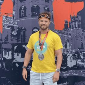 Stefan wearing a Lullaby Trust t-shirt and a medal, standing in front of a big tough mudder backdrop.