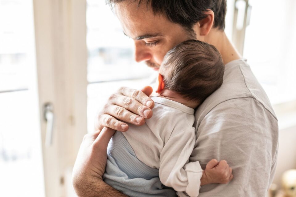 A young baby is resting against the shoulder of their father, who is pressing his head into the baby's and patting their back.