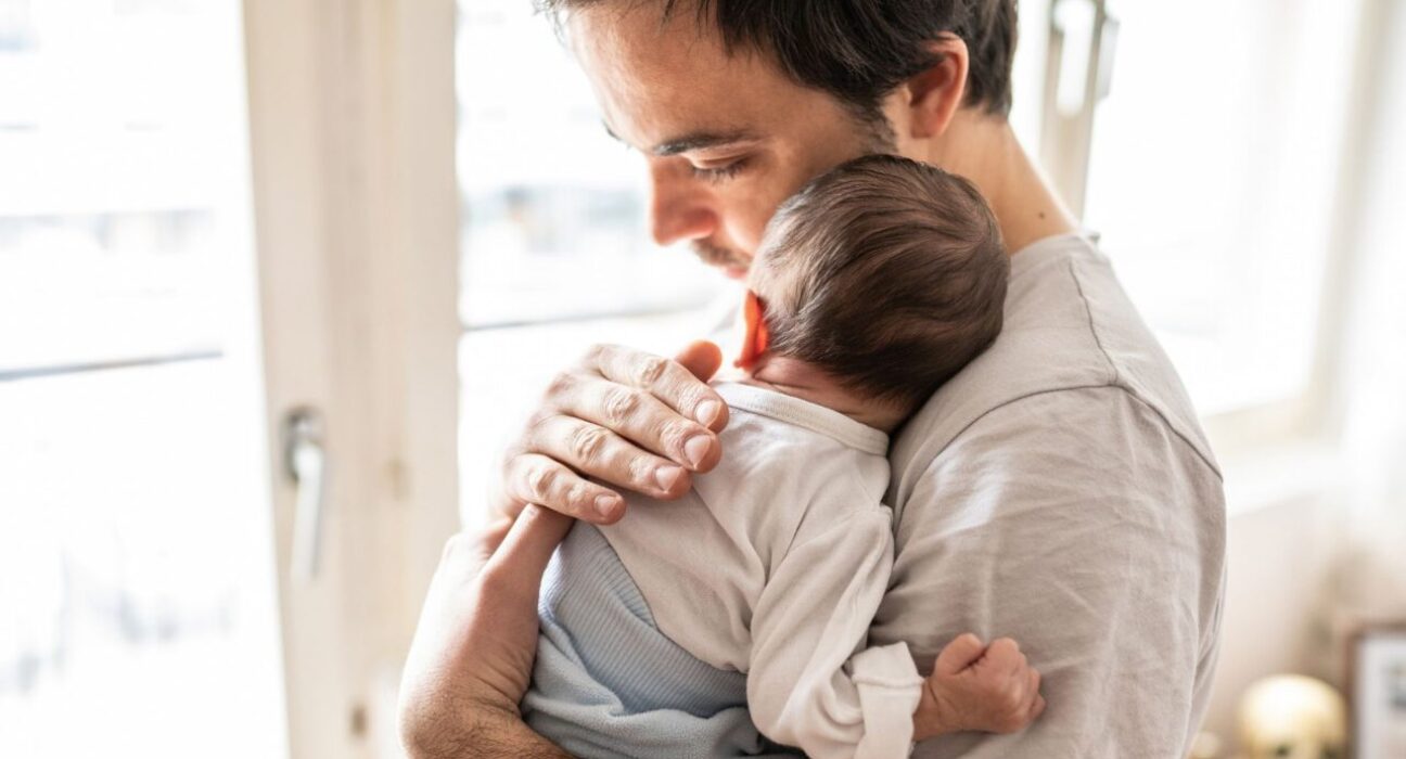 A young baby is resting against the shoulder of their father, who is pressing his head into the baby's and patting their back.