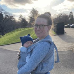 Laura and Sam are pictured outdoors. Laura is smiling with her hair in a bun, and Sam is strapped to her front, wearing a hat with teddy ears.