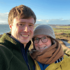 Matt and Laura are taking a selfie in front of green grassy moors. They are dressed warmly and both smiling.