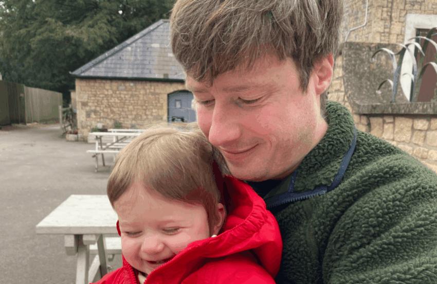 Matt and baby Sam are sat on a bench. Sam is grinning cheekily and Matt is holding him, smiling and looking down at baby Sam's hands.
