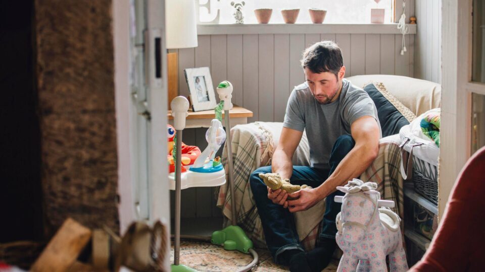 Man sitting on an armchair in a child's bedroom amongst toys, looking solemnly at a teddy bear in his hands.