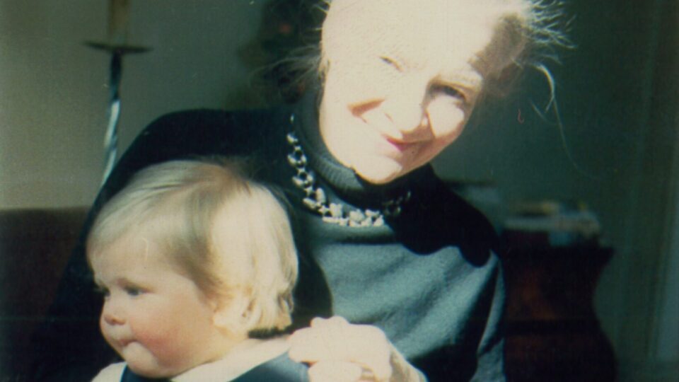 A lady holds a baby on her lap. She is smiling to the camera while the baby turns his head away.