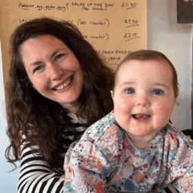 Beth sitting in a cafe wearing a stripy top, holding Poppy. Both are smiling at the camera.