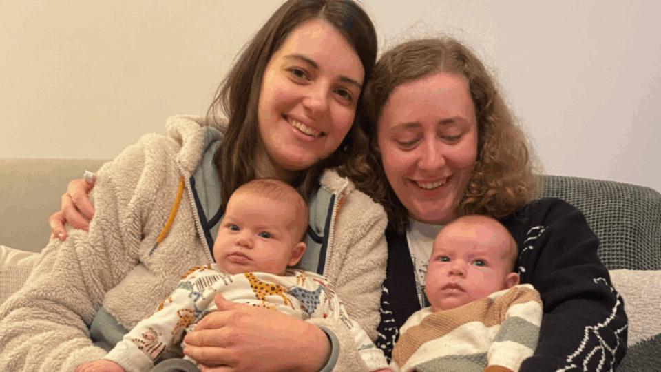 Izzy and Laura sitting next to each other on the sofa, holding Alo and Jovie.