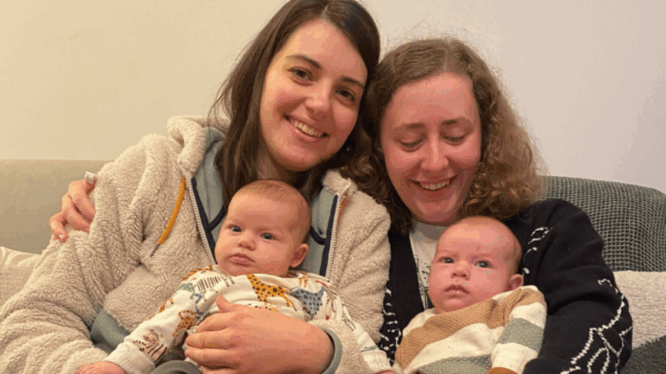 Izzy and Laura sitting next to each other on the sofa, holding Alo and Jovie.