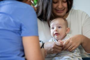 Parent sitting with her baby on her lap, in front of a health professional wearing scrubs whose back is towards the camera.