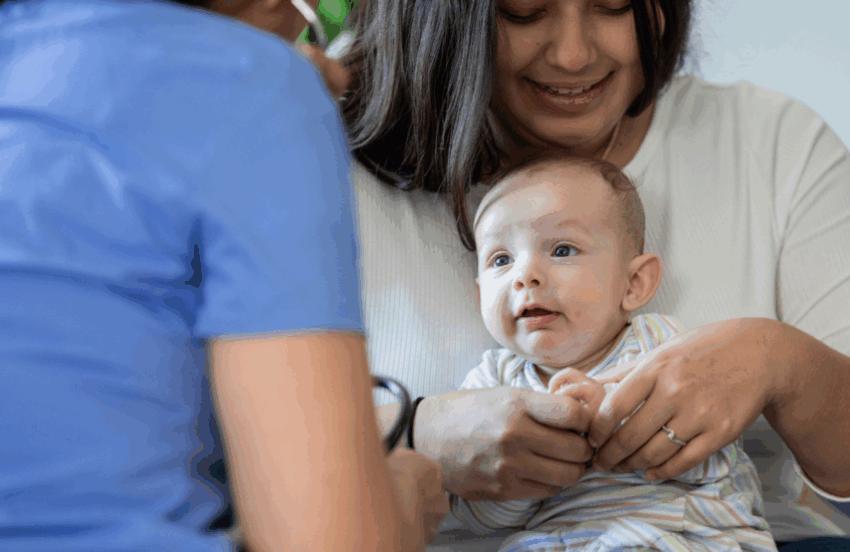 Parent sitting with her baby on her lap, in front of a health professional wearing scrubs whose back is towards the camera.