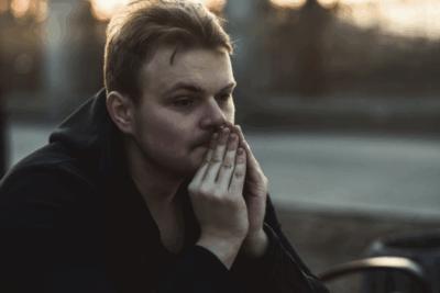 A man is looking into the distance with his hands up to his mouth. He looks deep in thought, and quite sad. The sun is setting behind him.