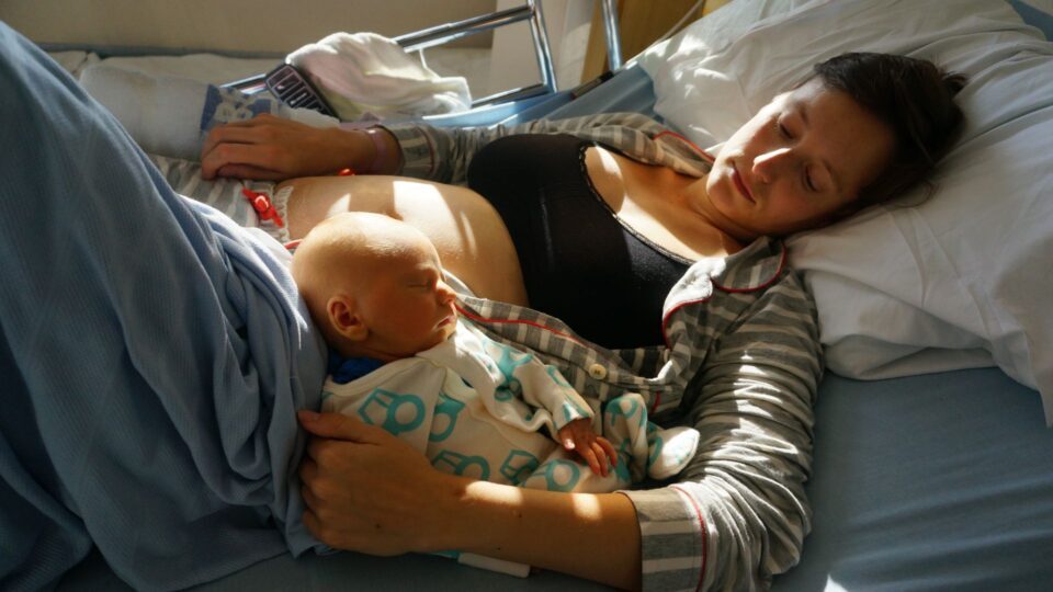 Kit and Sarah are lying on a hospital bed. Kit's head is resting on Sarah's stomach as he sleeps, and Sarah looks down at him.
