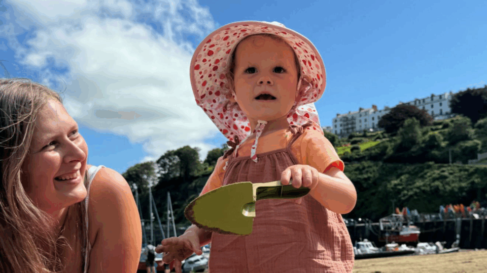 Olive and Gemma are at the beach on a sunny day, Olive has a sun hat on and is holding a spade.