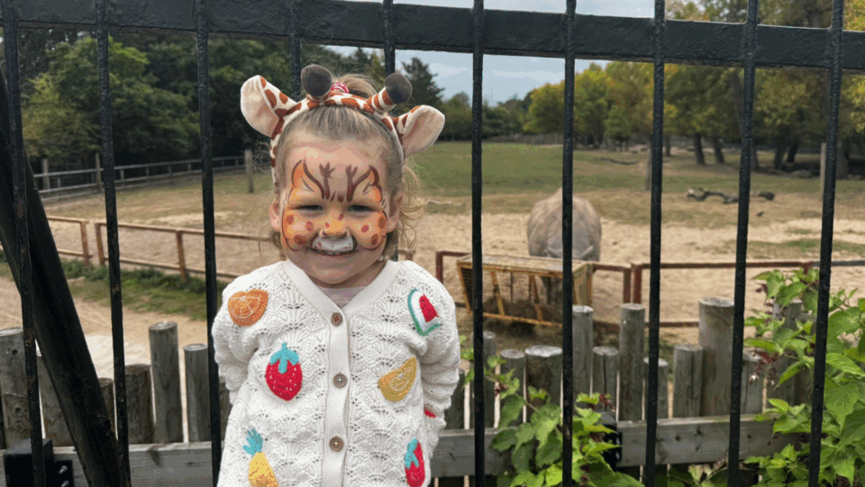 Olive has had her face painted like a giraffe and is stood by the rails of a zoo, behind a rhino