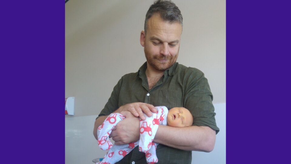 James is holding Kit on his arm. Kit is sleeping with his mouth open, and James smiles down at him.