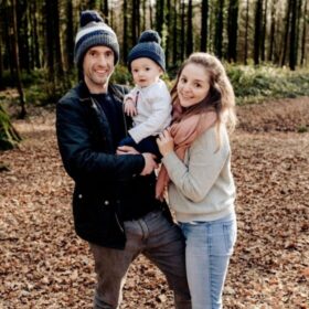 A man and woman hold their baby son while smiling to camera. They are standing in the woods, wearing winter clothing.