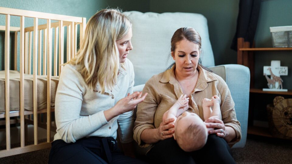 A mum is looking down at her baby in her knee. A health visitor is next to them, talking to the mother. A cot is in the background.