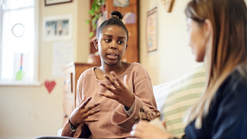 A woman is talking to a blurred woman on the couch. She is using hand gestures and making eye contact, she looks as though she is describing something difficult.