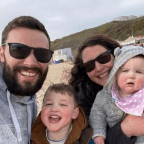 Beth and Callum with three-year-old Rory and eight-month-old Poppy. They are taking a selfie on a pebbly beach.