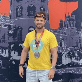 Stefan wearing a Lullaby Trust t-shirt and a medal, standing in front of a big tough mudder backdrop.
