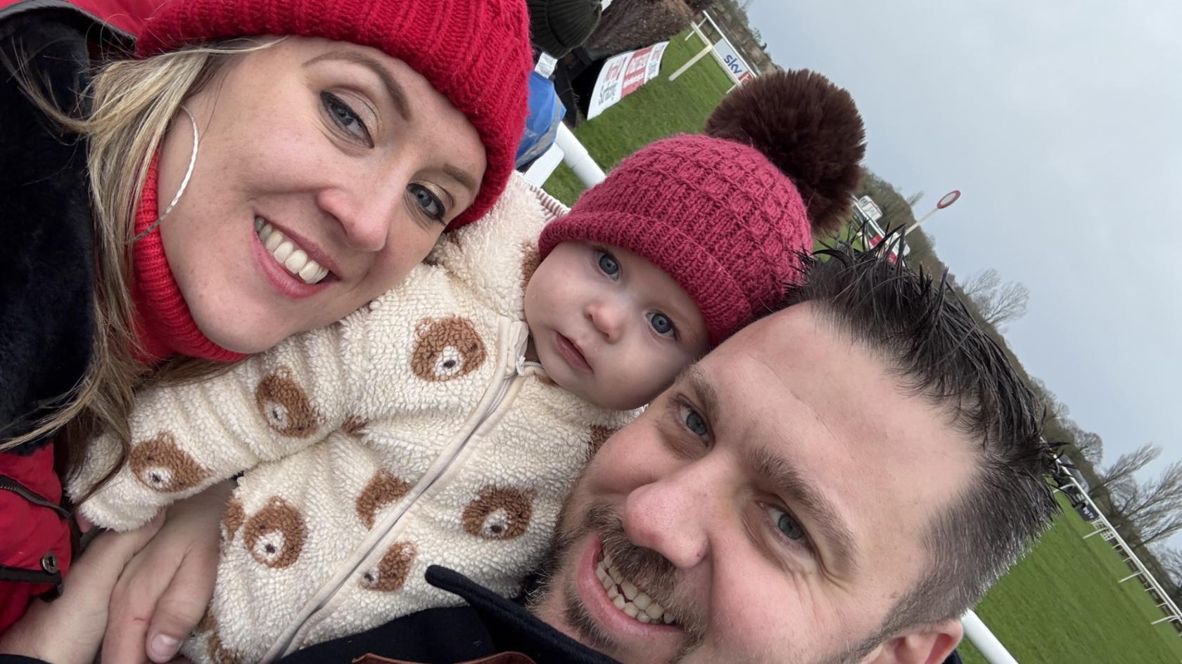 Helen wearing a red wooly hat, Faith also wearing a red hat, and Stephen, all smiling at the camera outdoors.