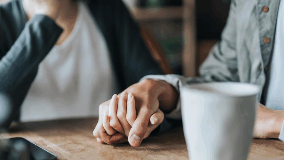 Two people are holding hands supportively. There is a phone and mug on the table.