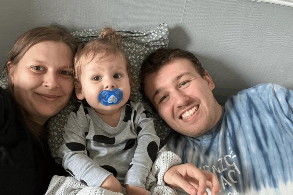 Gemma, Olive and Thomas are in bed together. Olive is inbetween Gemma and Olive and all are smiling. Olive has a dummy and messy bed hair.