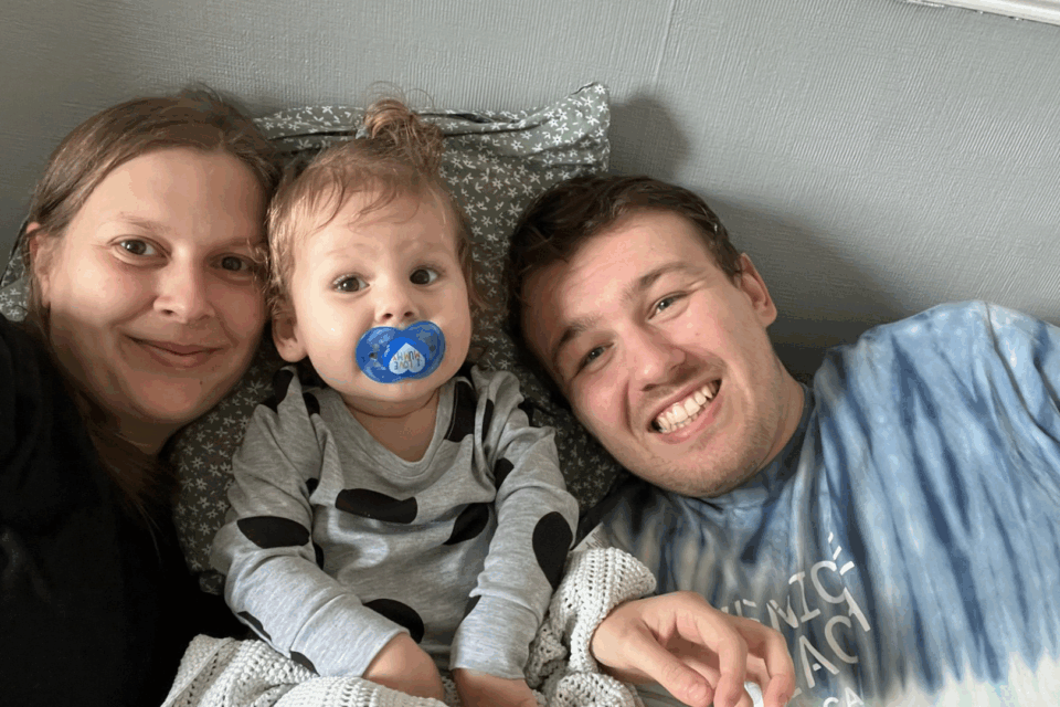Gemma, Olive and Thomas are in bed together. Olive is inbetween Gemma and Olive and all are smiling. Olive has a dummy and messy bed hair.