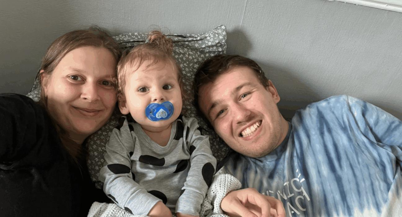 Gemma, Olive and Thomas are in bed together. Olive is inbetween Gemma and Olive and all are smiling. Olive has a dummy and messy bed hair.