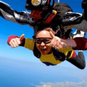Katie strapped to an instructor, in the middle of her skydive giving thumbs up to the camera.