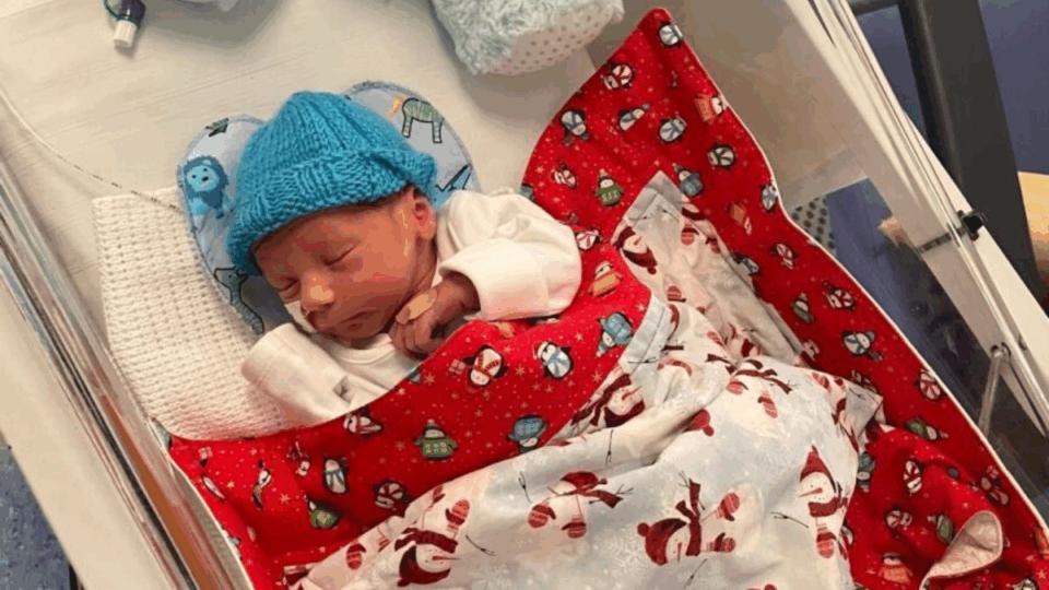 Baby George asleep in NICU with blanket on and a hat.