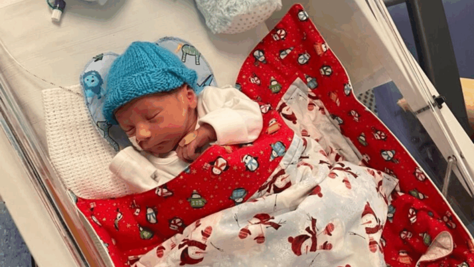 Baby George asleep in NICU with blanket on and a hat.
